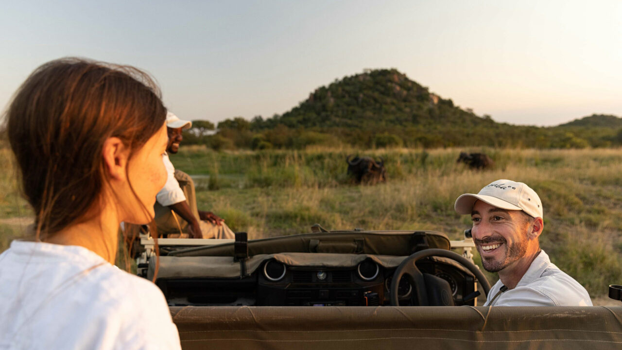 A guest and ranger share a laugh on a game drive, buffalo grazing beyond, Ulusaba, Sabi Sands, South Africa.