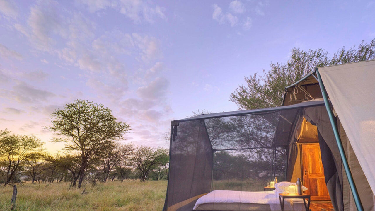 A safari tent glows against a dusk sky, its bed visible through the mesh screen, a mobile camp in the Serengeti, Tanzania.
