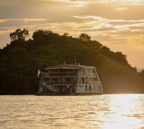 A multi-deck houseboat sits below a wooded hillside on glowing, golden water, Lake Kivu, Rwanda.