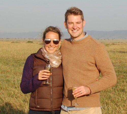 Smiling couple in jumpers hold drinks on open grassland with distant hills.
