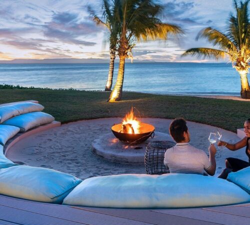 A couple on honeymoon safari raise wine glasses around a sunken fire pit on the beach at dusk, Miavana, Madagascar