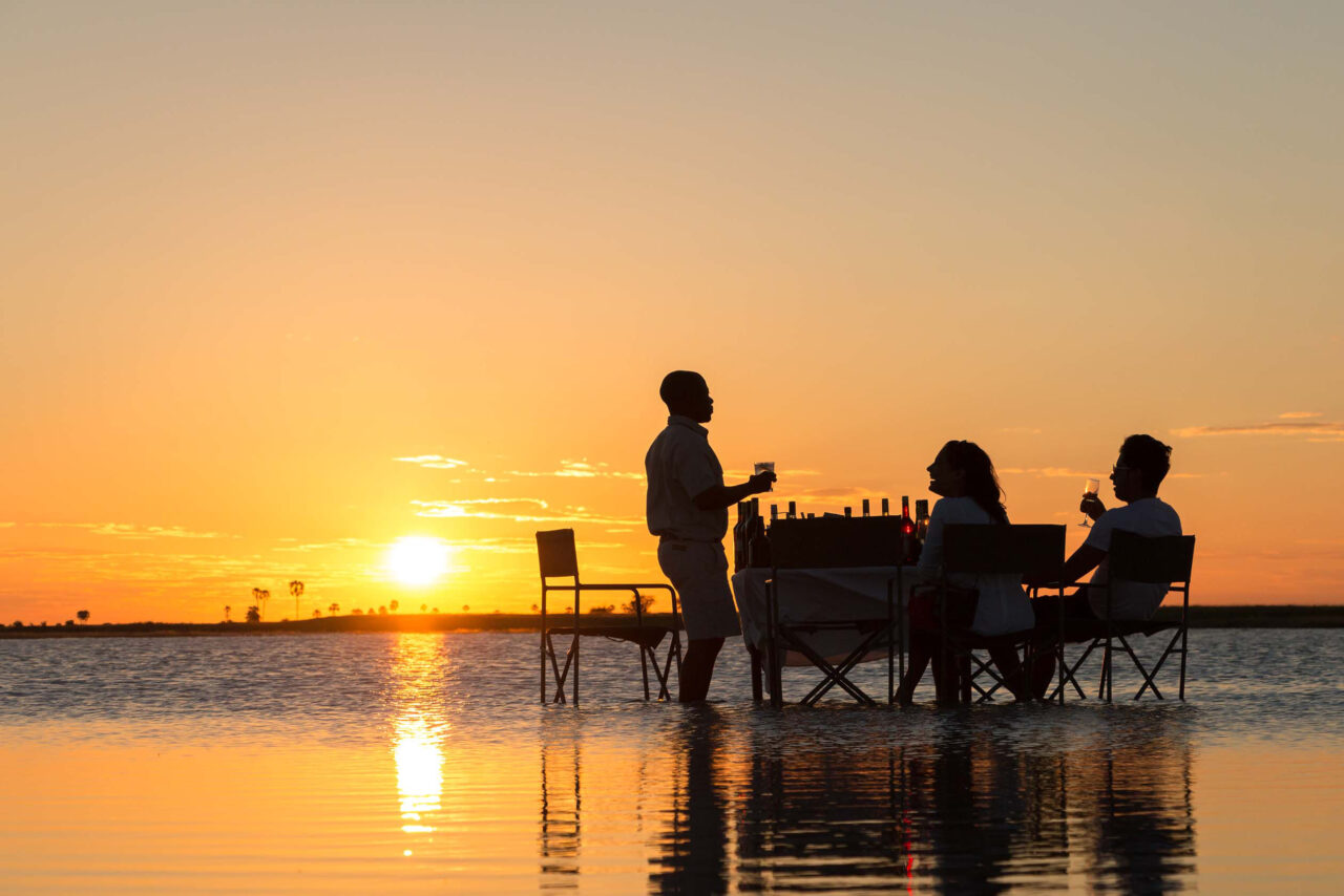 A couple at a dining table silhouetted against a golden sunset over the Makgadikgadi Salt Pans, Botswana, a stay at a luxury safari lodge.