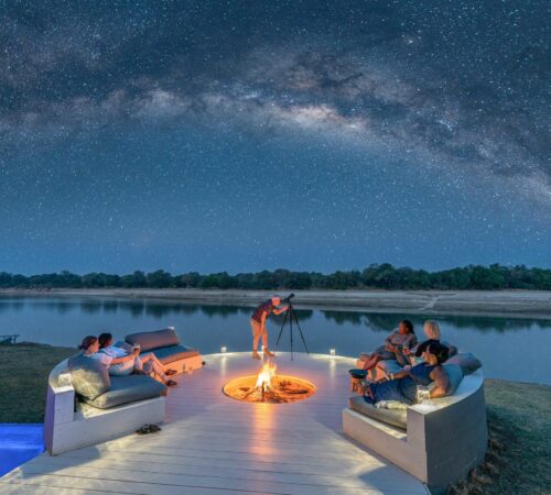 Guests relax around a fire on a riverside deck beneath the Milky Way, Chinzombo, South Luangwa, Zambia.