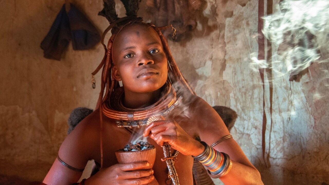 Himba woman wearing traditional ochre hairstyle, layered necklaces, and colorful bangles, holding a wooden vessel with smoking embers inside a clay-walled hut.
