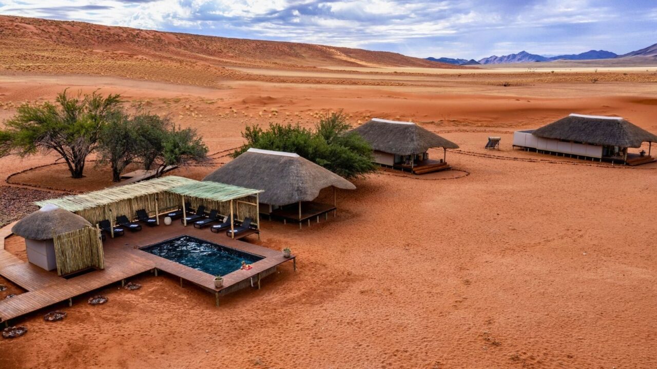 Thatched-roof desert lodges with a shaded outdoor pool sit amid the vast red sand dunes of a remote desert landscape, with scattered greenery providing contrast to the arid surroundings. The backdrop features rolling dunes and distant mountains under a partly cloudy sky.