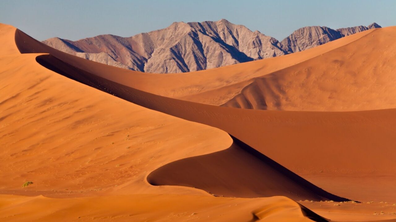 Sweeping view of towering orange sand dunes casting sharp shadows, with rugged gray mountains rising in the background under a clear blue sky.