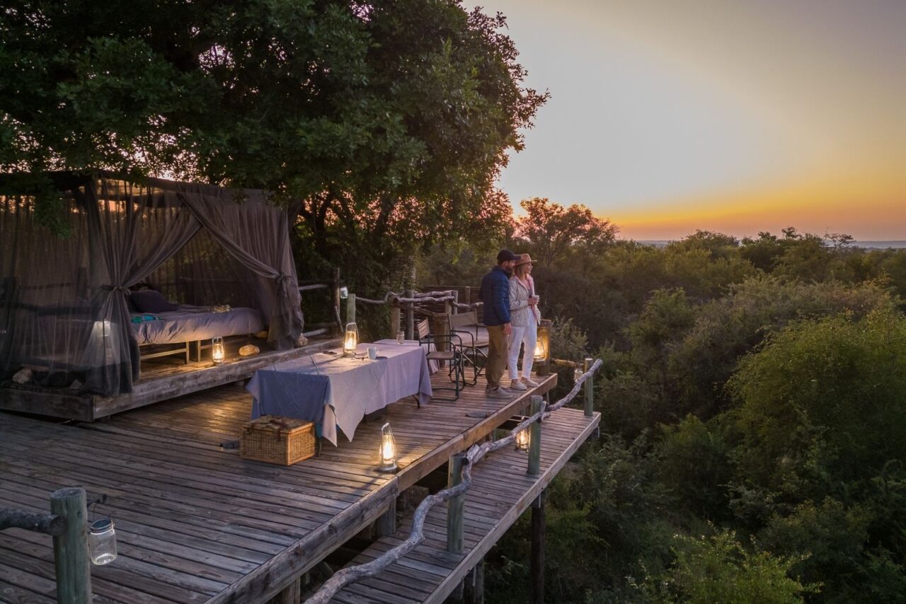 A couple on honeymoon safari watch the sunset from a sleep-out deck, netted bed and lantern-lit table, South Africa.